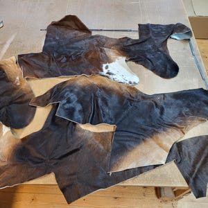 Può includere: Diversi pezzi irregolari di pelle bovina marrone, nera e bianca con una consistenza di pelliccia naturale sono disposti in piano. I pezzi presentano varie macchie marrone scuro e nere con occasionali segni bianchi.