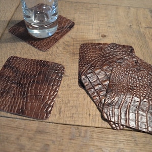 May include: Four brown leather coasters with a crocodile skin pattern. One coaster is on top of the other three.
