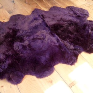 May include: A plush purple sheepskin rug with a soft, fluffy texture. The rug is laid out on a wooden floor.