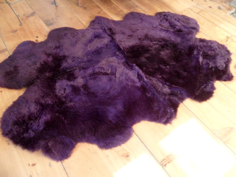 May include: A plush purple sheepskin rug with a soft, fluffy texture. The rug is laid out on a wooden floor.