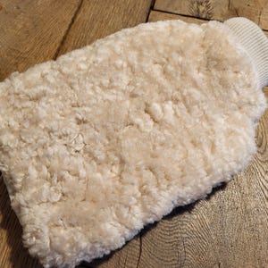 May include: Cream-colored, fluffy sheepskin mitt with a ribbed cuff. The mitt is rectangular with rounded edges, and the wool appears soft and dense. The background is a wooden surface.
