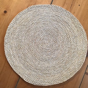 Könnte beinhalten: Ein runder, gewebter Platzteller aus Naturfasern. Der Platzteller hat eine hellbeige Farbe und ein kreisförmiges Muster.
