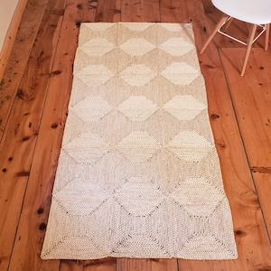 Op de afbeelding: Een rechthoekig vloerkleed met een herhalend ruitpatroon in beige en crèmetinten. Het kleed, gemaakt van natuurlijke vezels, ligt op een houten vloer. Het geometrische ontwerp voegt textuur toe. Ideaal voor een gang of kleine kamer.