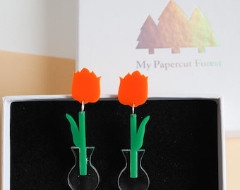 May include: A pair of acrylic tulip earrings. The earrings feature orange tulip blooms, green stems and leaves, and clear vase-shaped bases. The earrings are displayed in a white box with the text "My Papercut Forest".