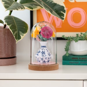 a paper vase with paper flowers inside in a plastic dome on a shelf. this is a kit by my papercut forest