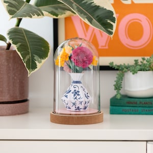 a paper vase with paper flowers inside in a plastic dome on a shelf. this is a kit by my papercut forest