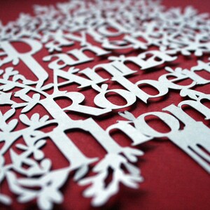 May include: A white paper cut-out of a tree with names, including "Robert" and "Anna", against a red background. The tree design features intricate leaf and branch details, creating a layered effect. The names are in a serif font.