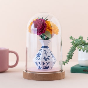 a paper vase with paper flowers inside in a plastic dome on a shelf. this is a kit by my papercut forest