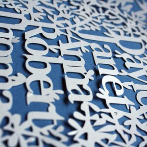 May include: A white paper cut-out design featuring names and floral elements against a blue background. The names are in a stylized font, and the overall design resembles a family tree or decorative artwork.