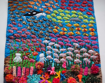 May include: A colorful textile art piece depicting an underwater scene. The artwork features a blue background with rows of embroidered fish in various colors, including orange, red, and yellow. Whales and coral are also present.