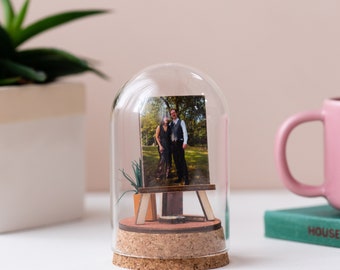 May include: A clear glass dome encases a small wooden easel displaying a photograph of a couple. The easel sits on a cork base with a small potted plant. A pink mug and a green book are in the background.