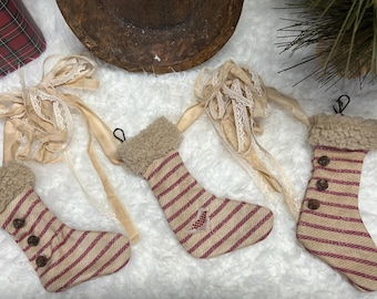 Puede incluir: Tres calcetines de Navidad a rayas rojas y blancas con ribete de piel sintética y botones marrones. Los calcetines están colgados de una superficie blanca y esponjosa.