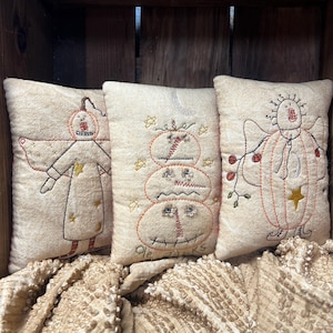 May include: Three beige decorative pillows with embroidered folk art designs. One pillow features a figure with a star and a red and yellow design. Another has a stacked pumpkin design with the text "Ole Tacks". The third pillow has a figure with a star.