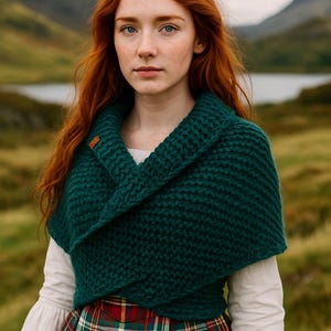 Pode incluir: Uma pessoa com cabelo ruivo e olhos azuis usa um xale de malha verde escuro e uma saia xadrez. O xale é preso com uma pequena etiqueta de couro castanho. O fundo mostra um lago e montanhas.