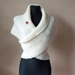 May include: Off-white, hand-knitted shawl with a wide collar and wrap-around style. The shawl is displayed on a black mannequin, secured with a small, brown leather tag. The knitted texture is visible.