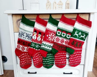 May include: Five Christmas stockings with red, white, and green patterns hang from a white dresser. Each stocking features a unique design, including snowflakes, reindeer, and snowmen. The stockings are made of knitted fabric and are ready for the holidays.