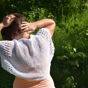 May include: A woman wearing a white crocheted top with a delicate lace pattern. The top is sleeveless and has a loose, flowing fit. The woman is standing in a grassy field.