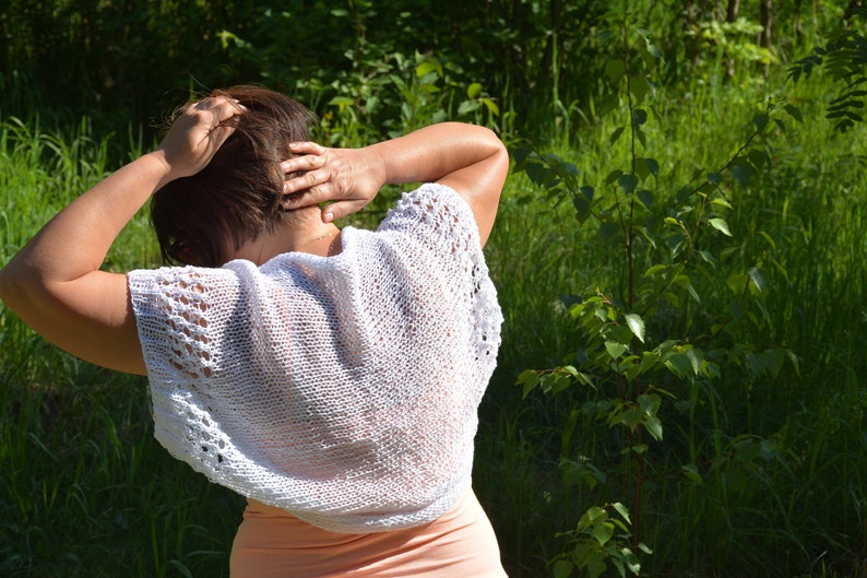 May include: A woman wearing a white crocheted top with a delicate lace pattern. The top is sleeveless and has a loose, flowing fit. The woman is standing in a grassy field.