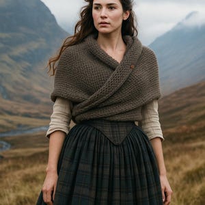 Puede incluir: Una mujer con un chal de punto marrón abrochado con un botón de madera. Lleva una camisa crema de manga larga y una falda a cuadros verde oscuro. El fondo muestra un paisaje montañoso.