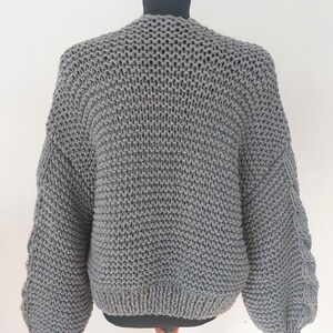 Pode incluir: Um cardigan cinza de malha grossa exibido em um manequim. O su&eacute;ter apresenta uma bainha e punhos largos e canelados, com um padr&atilde;o de malha texturizada no corpo e nas mangas. As mangas t&ecirc;m um design &uacute;nico, ligeiramente bufante. O cardigan &eacute; de cor neutra.