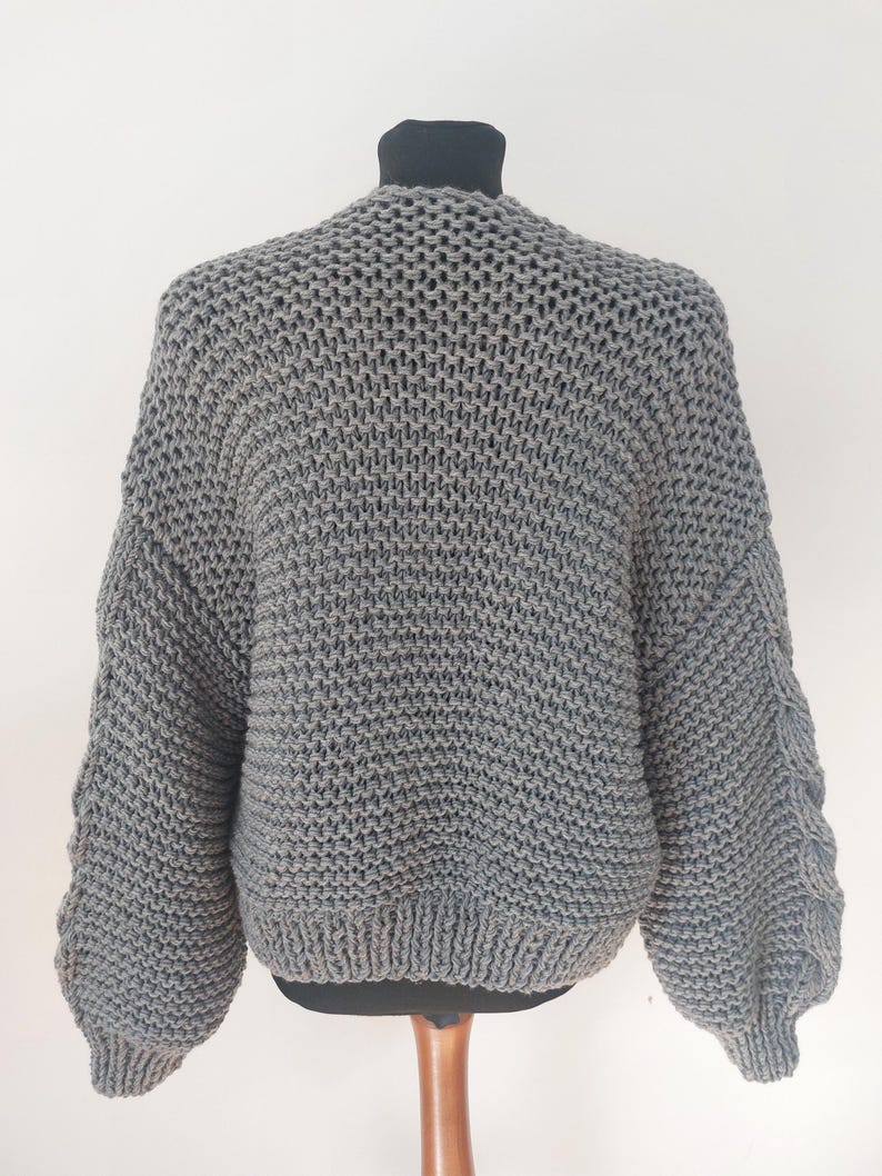 Pode incluir: Um cardigan cinza de malha grossa exibido em um manequim. O su&eacute;ter apresenta uma bainha e punhos largos e canelados, com um padr&atilde;o de malha texturizada no corpo e nas mangas. As mangas t&ecirc;m um design &uacute;nico, ligeiramente bufante. O cardigan &eacute; de cor neutra.