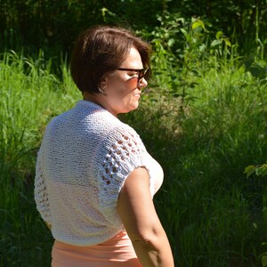 May include: A white crocheted shrug with a delicate openwork pattern. The shrug has short sleeves and a loose fit.