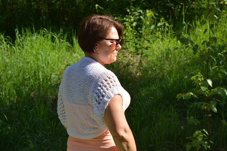 May include: A white crocheted shrug with a delicate openwork pattern. The shrug has short sleeves and a loose fit.