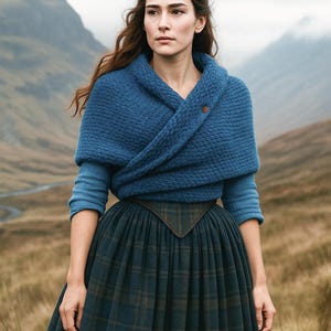 Könnte beinhalten: Eine Frau trägt einen blauen Strickschal mit Knopf, über einem langärmligen Oberteil. Sie trägt auch einen langen, plissierten karierten Rock. Der Hintergrund zeigt eine bergige Landschaft und einen bewölkten Himmel.