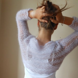 May include: A light purple knitted shrug with a delicate lace pattern. The shrug has long sleeves and a loose fit.