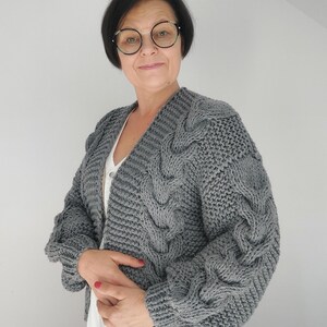 Pode incluir: Um cardigan cinza de malha grossa com um design de malha de cabo nas mangas e na frente. O cardigan tem decote em V e mangas compridas. A mulher na imagem est&aacute; usando o cardigan sobre um top branco e uma saia branca.