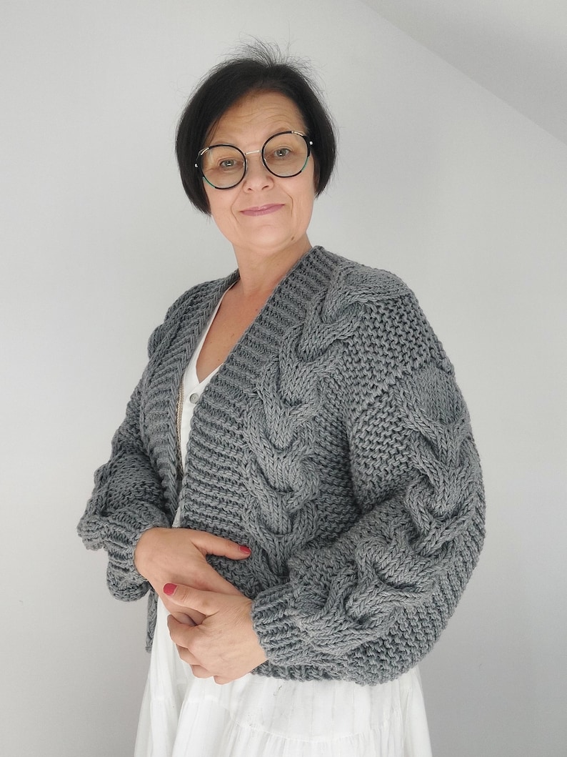 Pode incluir: Um cardigan cinza de malha grossa com um design de malha de cabo nas mangas e na frente. O cardigan tem decote em V e mangas compridas. A mulher na imagem est&aacute; usando o cardigan sobre um top branco e uma saia branca.