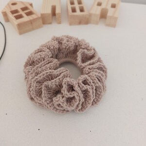 May include: A light brown, crocheted scrunchie with a ruffled design. The hair accessory is circular and made of a soft, textured yarn. In the background are small, wooden house-shaped decorations.