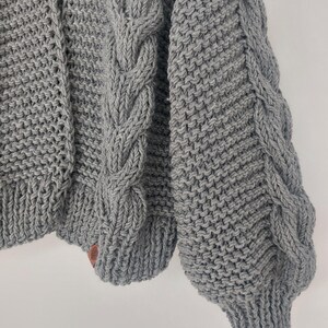 Pode incluir: Um close-up de um cardigan cinza tricotado. O cardigan apresenta um design de malha de cabo nas mangas e pain&eacute;is frontais. As mangas t&ecirc;m um punho canelado. Uma pequena etiqueta de couro marrom &eacute; costurada na parte inferior do cardigan.