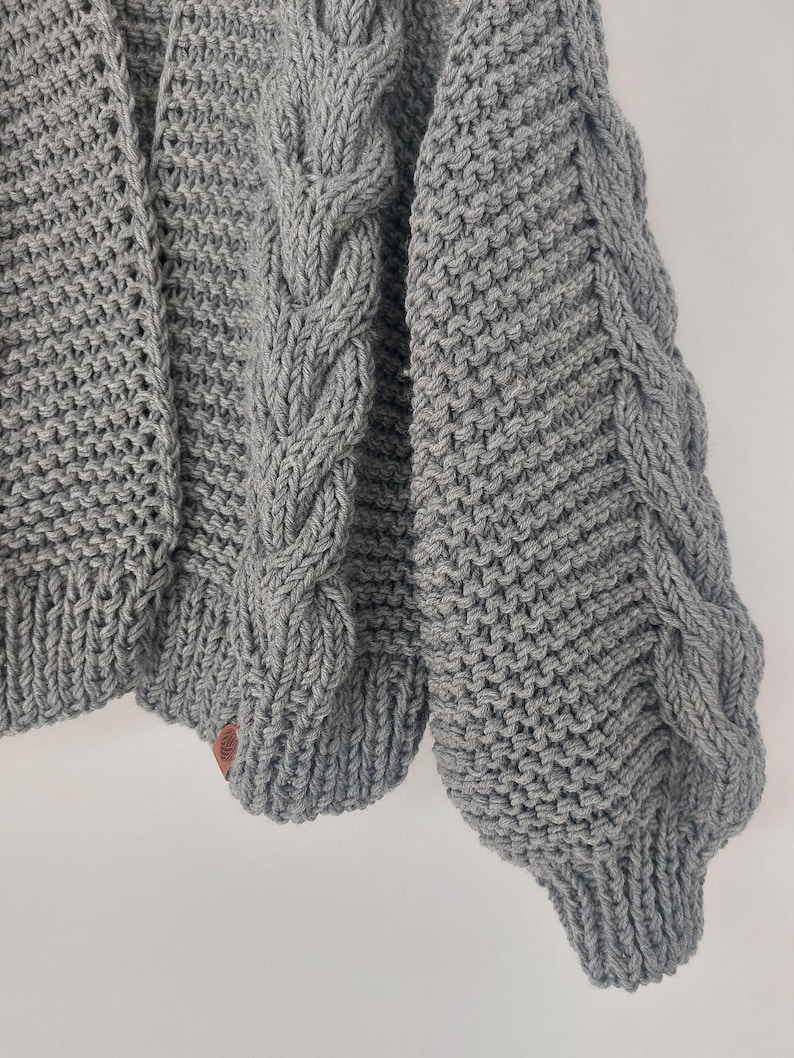 Pode incluir: Um close-up de um cardigan cinza tricotado. O cardigan apresenta um design de malha de cabo nas mangas e pain&eacute;is frontais. As mangas t&ecirc;m um punho canelado. Uma pequena etiqueta de couro marrom &eacute; costurada na parte inferior do cardigan.