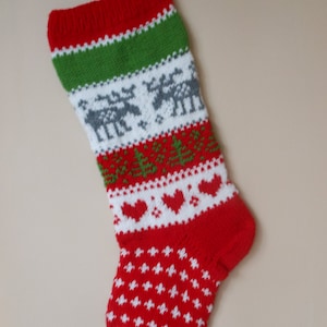 Può includere: Un calzino di Natale rosso e verde a maglia con sfondo bianco. Il calzino presenta un motivo di renne, cuori e fiocchi di neve.
