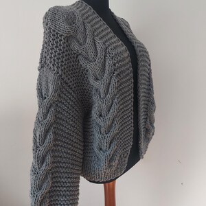 Pode incluir: Um cardigan cinza de malha grossa com um design de malha de cabo nas mangas e na frente. O su&eacute;ter aberto &eacute; exibido em um manequim preto, mostrando seu ajuste texturizado e grande. As mangas t&ecirc;m formato de bal&atilde;o.