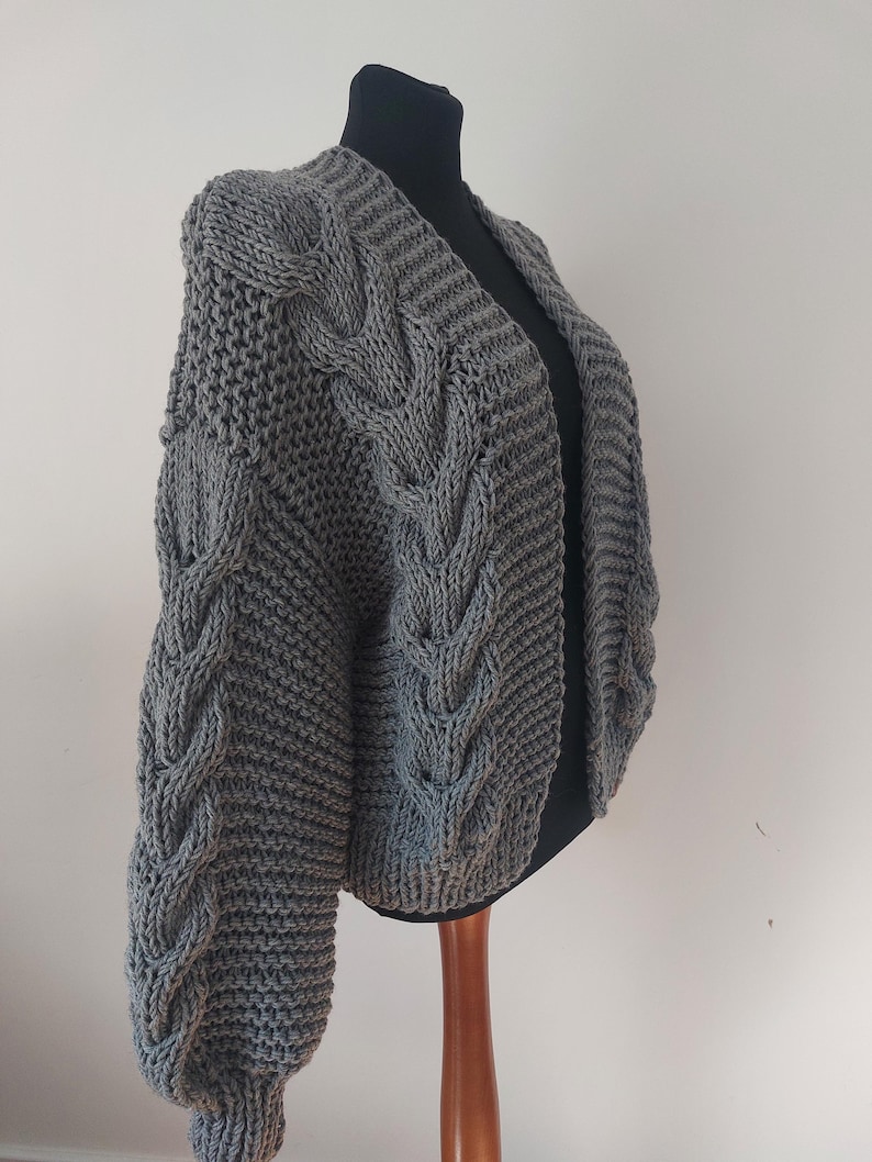 Pode incluir: Um cardigan cinza de malha grossa com um design de malha de cabo nas mangas e na frente. O su&eacute;ter aberto &eacute; exibido em um manequim preto, mostrando seu ajuste texturizado e grande. As mangas t&ecirc;m formato de bal&atilde;o.