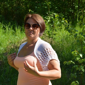 May include: A woman wearing a white crocheted shrug over a peach tank top and sunglasses. She is standing in a grassy area with green foliage.