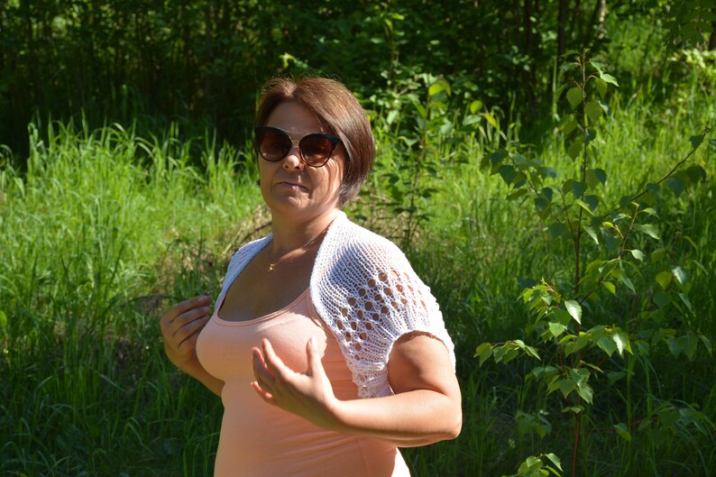 May include: A woman wearing a white crocheted shrug over a peach tank top and sunglasses. She is standing in a grassy area with green foliage.