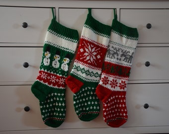 May include: Three knitted Christmas stockings hanging in a row. The left stocking features snowmen, the middle has a large snowflake pattern, and the right displays reindeer. All three stockings incorporate red, green, white, and grey yarn colors.