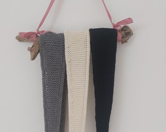 May include: Three knitted wall hangings in grey, cream, and black, suspended from a natural wood branch with a red and white ribbon. The knitted pieces have a long, triangular shape, and are displayed against a white wall.