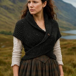 May include: A dark grey knitted wrap with a button fastening. The wrap is worn over a cream-coloured long-sleeved shirt and a brown plaid skirt. The background shows a mountain and a loch.