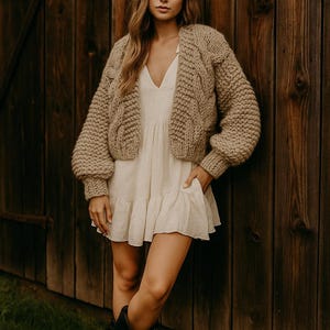 Peut inclure: Une femme porte un cardigan beige en tricot torsadé sur une robe blanche à ourlet volanté. La tenue est complétée par des bottes de cowboy noires. L'arrière-plan est un mur en bois.