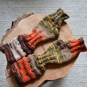 May include: Two pairs of knitted arm warmers with a striped pattern in shades of brown, orange, green, and yellow. The arm warmers are laid out on a wooden surface.