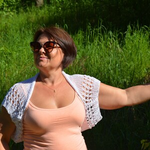May include: A woman wearing a white crocheted shrug over a peach tank top. She is wearing sunglasses and has short brown hair. She is standing in a grassy area with a tree branch in the background.