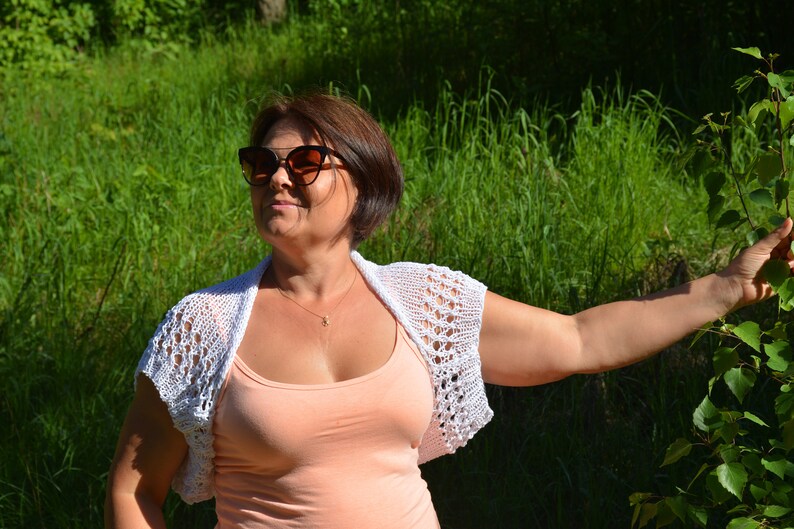 May include: A woman wearing a white crocheted shrug over a peach tank top. She is wearing sunglasses and has short brown hair. She is standing in a grassy area with a tree branch in the background.