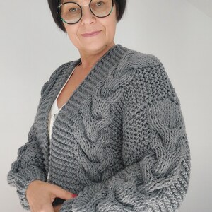 Pode incluir: Um cardigan cinza de malha com gola em V. O su&eacute;ter apresenta uma malha grossa e texturizada com intrincados padr&otilde;es de cabo nas mangas e pain&eacute;is frontais. O cardigan &eacute; de cor cinza s&oacute;lida e tem um ajuste folgado.