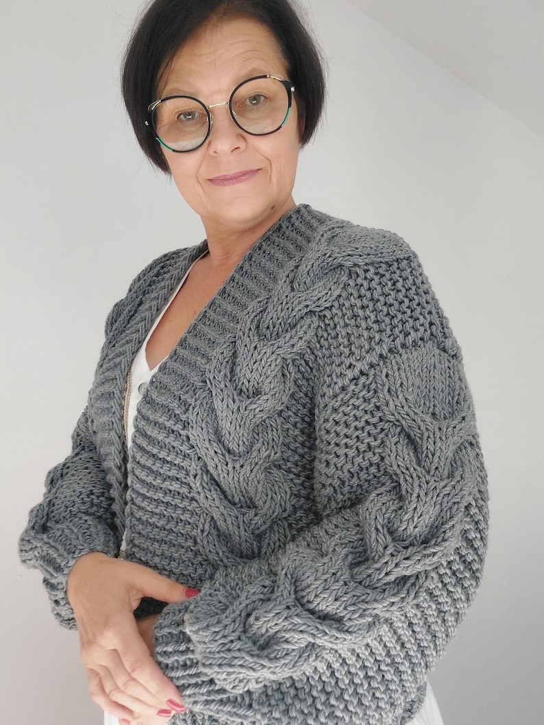 Pode incluir: Um cardigan cinza de malha com gola em V. O su&eacute;ter apresenta uma malha grossa e texturizada com intrincados padr&otilde;es de cabo nas mangas e pain&eacute;is frontais. O cardigan &eacute; de cor cinza s&oacute;lida e tem um ajuste folgado.