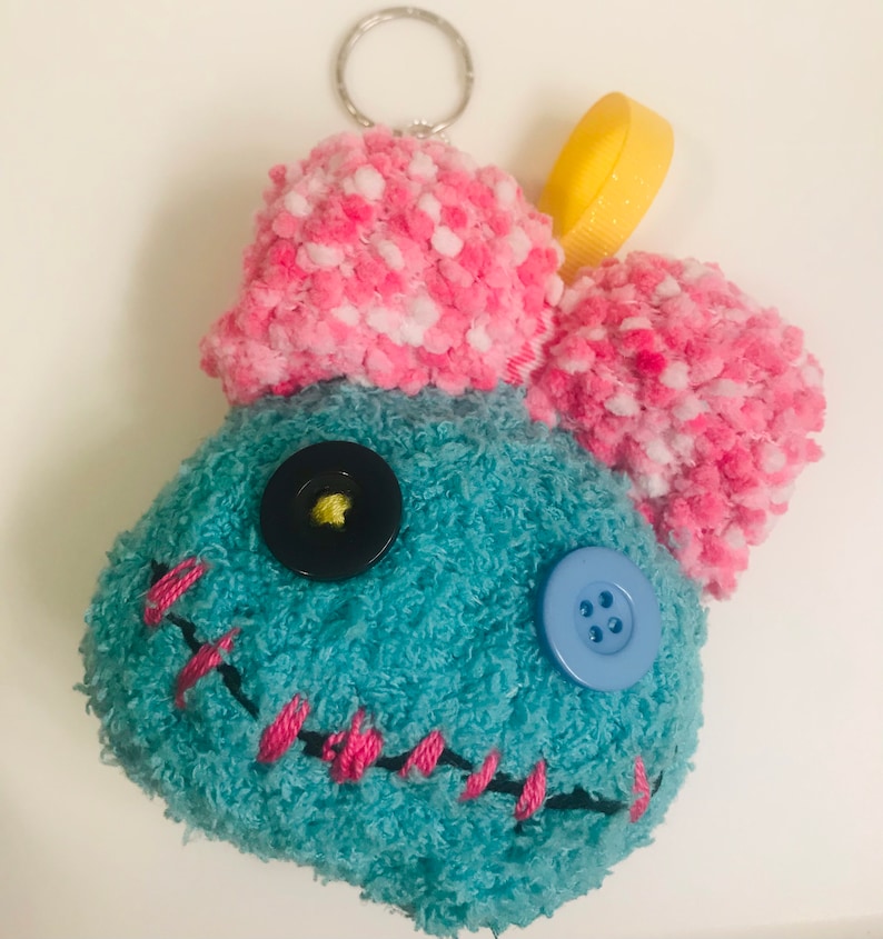 Pu&ograve; includere: Portachiavi in ​​peluche fatto a mano a forma di personaggio dei cartoni animati. Il personaggio &egrave; verde acqua con cuciture rosa e una parte superiore a pompon rosa e bianca. Ha un bottone nero e uno blu per gli occhi e un nastro giallo.