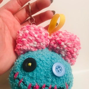 Pu&ograve; includere: Portachiavi in ​​peluche fatto a mano a forma di personaggio dei cartoni animati. Il personaggio ha un corpo blu con cuciture rosa, un fiocco rosa e bianco e due occhi a bottone. Sono attaccati un nastro giallo e un portachiavi argentato.